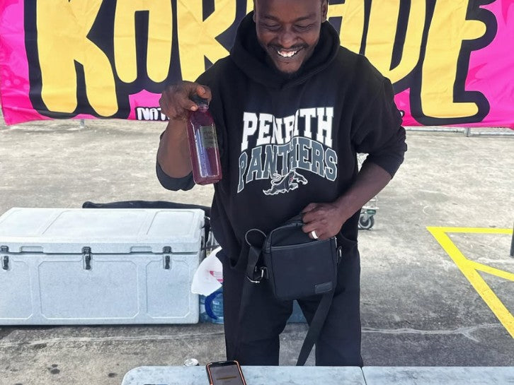 Mohamed holding a bottle of Karkade in front of the Karkade banner at Westfield Mt Druitt